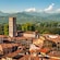 A scenic view featuring historic buildings with terracotta rooftops, a tall bell tower and a backdrop of mountains